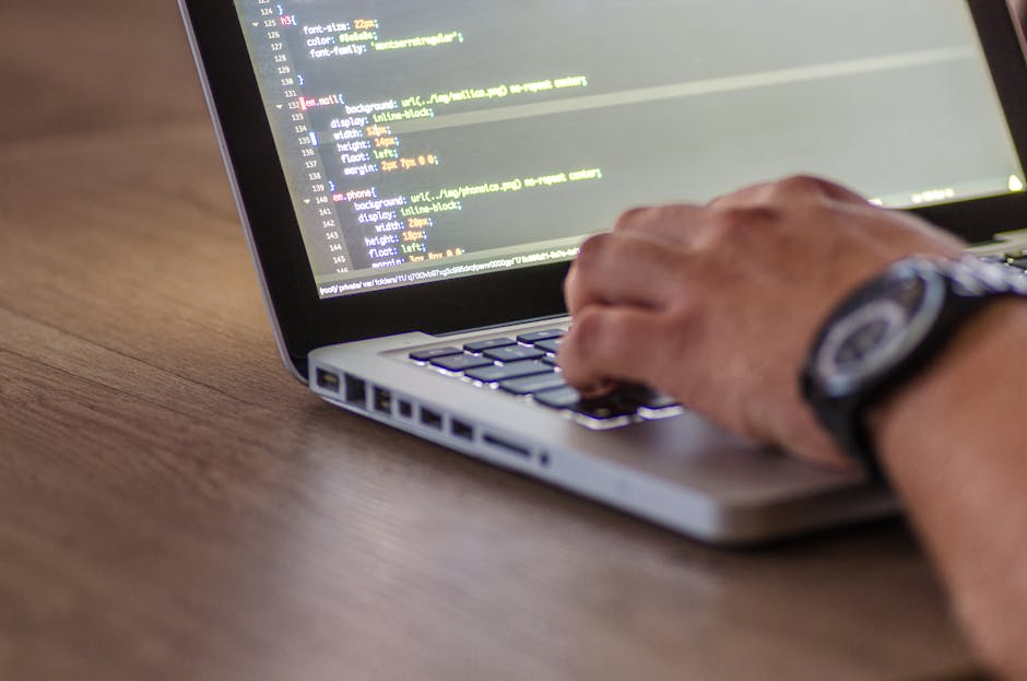 A close-up of a person's hand typing on a laptop keyboard placed on a wooden desk. The laptop screen displays lines of colorful code, indicating programming or web development activity. The person is